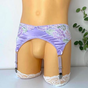 Op de afbeelding: Een lila satijnen jarretelgordel met bloemenkanten afwerking in paarse, groene en witte tinten. De jarretelgordel heeft verstelbare bandjes en metalen clips. De onderkant van de jarretelgordel heeft witte kanten afwerking.