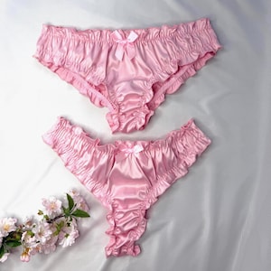 Può includere: Due paia di biancheria intima in raso rosa chiaro con bordi arricciati lungo la vita e le aperture delle gambe. Ogni paio è decorato con un piccolo fiocco in raso abbinato centrato sulla fascia in vita anteriore. Il tessuto ha una finitura liscia e lucida.