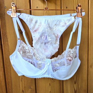 Può includere: Un completo intimo bianco, composto da un reggiseno e slip abbinati, è appeso a un gancio color oro rosa. Il reggiseno presenta delicati dettagli in pizzo e un piccolo fiocco. Lo slip ha un motivo floreale dai colori pastello.