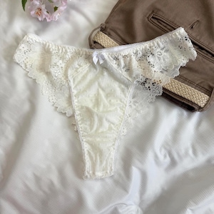 Può includere: Uno slip in pizzo color crema caratterizzato da un delicato motivo floreale lungo i bordi e da un piccolo fiocco bianco al centro. Il pannello centrale è realizzato in un tessuto trasparente a pois, che dona consistenza al capo.