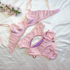 Può includere: Un completo intimo rosa chiaro composto da un reggiseno con ferretto in raso e pizzo con spalline regolabili e slip coordinati rifiniti in pizzo, dotati di reggicalze con clip metalliche e decorati con fiori artificiali.