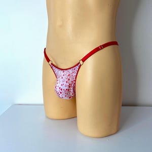 May include: A white and red heart-patterned thong with red straps and gold-coloured rings. The thong is displayed on a mannequin. The garment is designed for intimate apparel.
