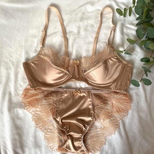 May include: A champagne-coloured lingerie set. The bra has underwire support and delicate lace trim on the cups. The matching briefs feature similar lace detailing around the edges. The set is made of a smooth, satin-like material, and is designed for comfort and style.