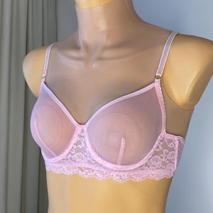 Könnte beinhalten: Ein hellrosa BH mit transparenten Cups und zarter Spitze am unteren Rand. Der BH hat verstellbare Träger und wird auf einer Schaufensterpuppe präsentiert. Das Design ist feminin und elegant.