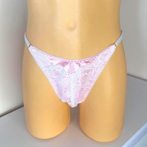 Könnte beinhalten: Rosafarbener Satin-String-Tanga mit weißen Spitzendetails. Der String hat eine zarte Spitzenborte an den Rändern und einen dünnen, hellblauen Gummibund. Die Vorderseite ist mit aufwendigen Spitzendesigns verziert.
