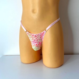 Puede incluir: Un tanga con un estampado de corazones rosas y blancos. Tiene una cinturilla rosa claro y tirantes finos. La tela está cubierta de pequeños corazones rosas y rojos. El tanga se exhibe en un maniquí.