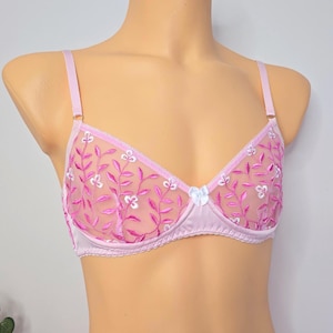 Op de afbeelding: Een lichtpaarse bh met transparante mesh cups voorzien van felroze geborduurde rank- en bladpatronen, geaccentueerd met kleine witte bloemdetails. Het kledingstuk is voorzien van verstelbare schouderbandjes en een klein wit strikje in het midden.