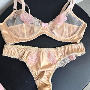 Può includere: Un completo di lingerie beige composto da un reggiseno e un perizoma abbinato. Il reggiseno presenta coppe in rete trasparente con ricami floreali rosa, un piccolo fiocco centrale rosa e spalline regolabili per la vestibilità.