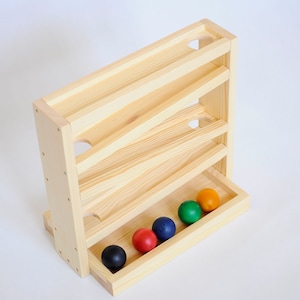 Könnte beinhalten: Holzspielzeug mit einer Rampe und fünf bunten Bällen. Die Rampe hat drei Ebenen und die Bälle sind schwarz, rot, blau, grün und orange.