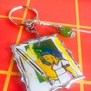 May include: A handmade keychain featuring a colorful illustration of a person with a sword. The artwork is set against a yellow and green background, framed with a silver border. The keychain includes a silver keyring and a decorative green bead.