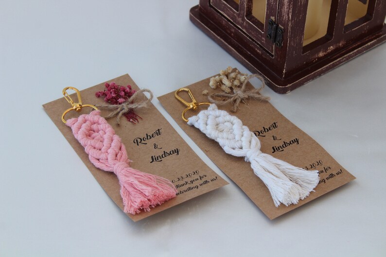 Puede incluir: Dos llaveros de regalo de boda con un dise&ntilde;o de macram&eacute;. El llavero de la izquierda es rosa y el de la derecha es blanco. Ambos llaveros tienen un llavero dorado y un borla. La tarjeta adjunta a cada llavero dice "Robert & Lindsay" e incluye la fecha de la boda.