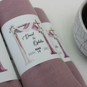 May include: Two dusty rose fabric squares with white labels attached. The labels feature a watercolor illustration of a wooden archway decorated with white and pink fabric, flowers, and greenery. The text on the labels reads "Daniel & Ophelia, Saturday MAR 19, 2022".