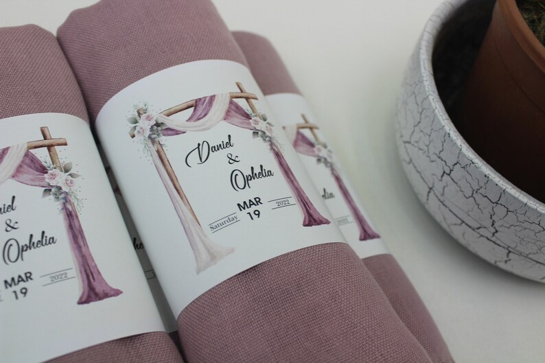 May include: Two dusty rose fabric squares with white labels attached. The labels feature a watercolor illustration of a wooden archway decorated with white and pink fabric, flowers, and greenery. The text on the labels reads "Daniel & Ophelia, Saturday MAR 19, 2022".