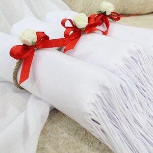 May include: Three white scarves with red bows and white flowers tied around them. The scarves are folded and arranged on a white and brown surface.