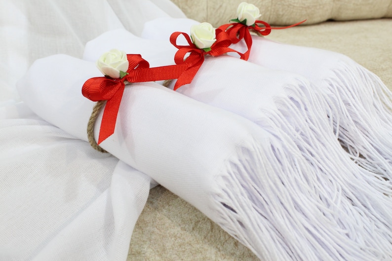 May include: Three white scarves with red bows and white flowers tied around them. The scarves are folded and arranged on a white and brown surface.