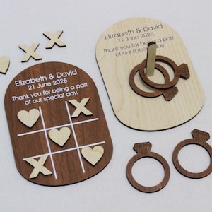 May include: A wooden wedding games set including a tic-tac-toe board, ring toss game, and diamond ring cutouts. The tic-tac-toe board has hearts and X's. The ring toss game has two rings. The text reads "Elizabeth & David 21 June 2025".