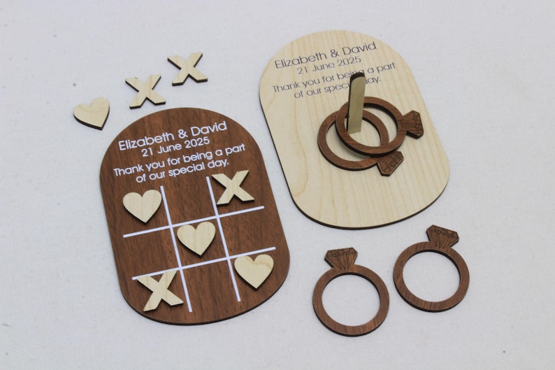 K&ouml;nnte beinhalten: Holzhochzeitsspiel-Set mit einem Tic-Tac-Toe-Brett, einem Ringwurfspiel und Diamantring-Ausschnitten. Das Tic-Tac-Toe-Brett hat Herzen und X's. Das Ringwurfspiel hat zwei Ringe. Der Text lautet "Elizabeth & David 21 June 2025".