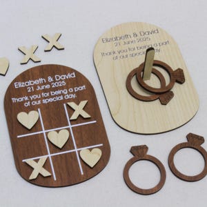 May include: Wooden wedding favors featuring a tic-tac-toe game, ring toss, and ring-shaped decorations. The tic-tac-toe board is brown with white lines and wooden X's and hearts. The ring toss game is on a light wood base. Text reads "Elizabeth & David 21 June 2025".