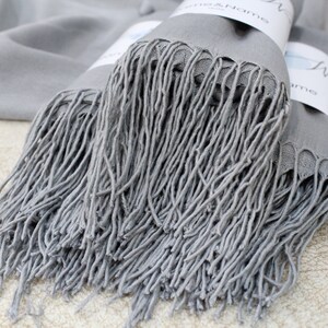 May include: A gray pashmina scarf with long fringe. The scarf is folded and stacked on a light brown surface.