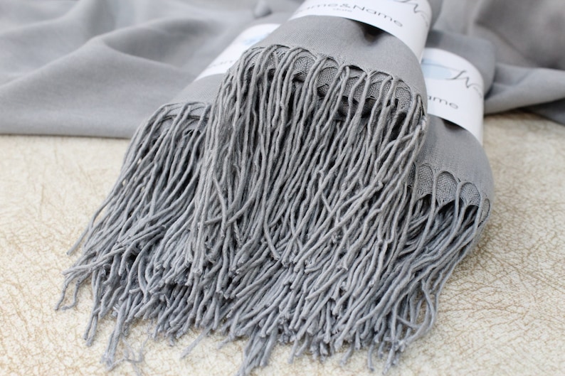May include: A gray pashmina scarf with long fringe. The scarf is folded and stacked on a light brown surface.