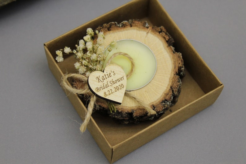 Op de afbeelding: Een kleine, ronde houten kaarsenhouder met een witte kaars erin. De houder is versierd met gedroogde witte bloemen en een hartvormig houten label met de tekst "Katie's Bridal Shower 8.22.2020". De houder zit in een bruine kartonnen doos.