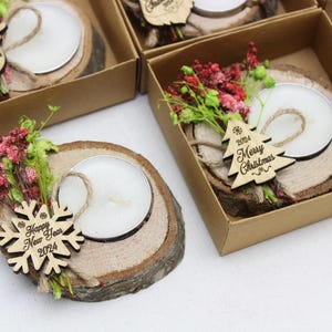 May include: A set of four small wooden candle holders, each containing a tea light candle. Each holder is decorated with a small wooden ornament, a sprig of dried flowers, and a piece of twine. The ornaments have the text "Happy New Year 2024" and "Merry Christmas 2024" on them.
