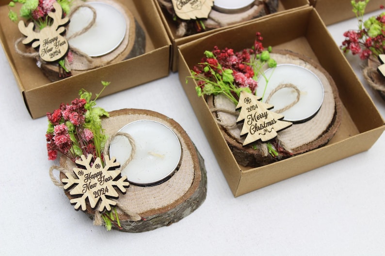 May include: A set of four small wooden candle holders, each containing a tea light candle. Each holder is decorated with a small wooden ornament, a sprig of dried flowers, and a piece of twine. The ornaments have the text "Happy New Year 2024" and "Merry Christmas 2024" on them.