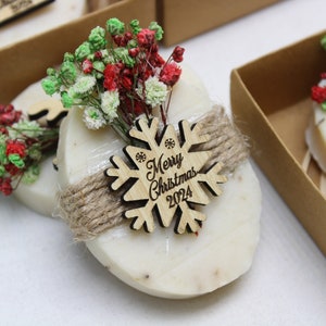 May include: Oval-shaped soap bars with a decorative wooden snowflake that reads "Merry Christmas 2024". Each bar is wrapped with twine and adorned with red and green dried flowers. The soaps are presented in small brown cardboard boxes.