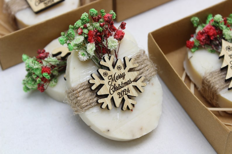 May include: Oval-shaped soap bars with a decorative wooden snowflake that reads "Merry Christmas 2024". Each bar is wrapped with twine and adorned with red and green dried flowers. The soaps are presented in small brown cardboard boxes.