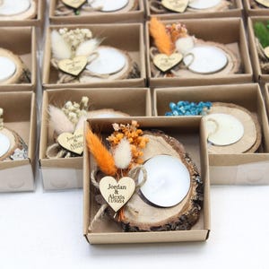 May include: Small gift boxes with a rustic theme. Each box contains a wooden slice with a white candle, dried flowers, and a wooden heart with a name and date. The boxes are arranged on a white surface.