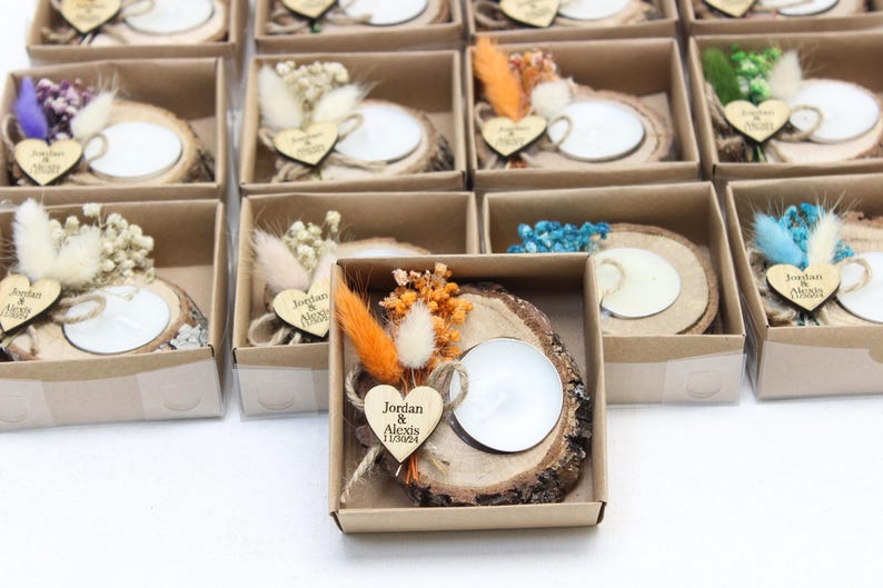 May include: Small gift boxes with a rustic theme. Each box contains a wooden slice with a white candle, dried flowers, and a wooden heart with a name and date. The boxes are arranged on a white surface.