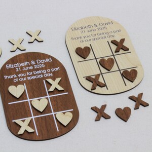 K&ouml;nnte beinhalten: Holz-Tic-Tac-Toe-Spielgeschenke mit graviertem Text. Ein Spielbrett ist dunkelbraun, das andere helles Holz. Die Spielsteine umfassen Holzherzen und X's. Der Text lautet "Elizabeth & David 21. Juni 2025".