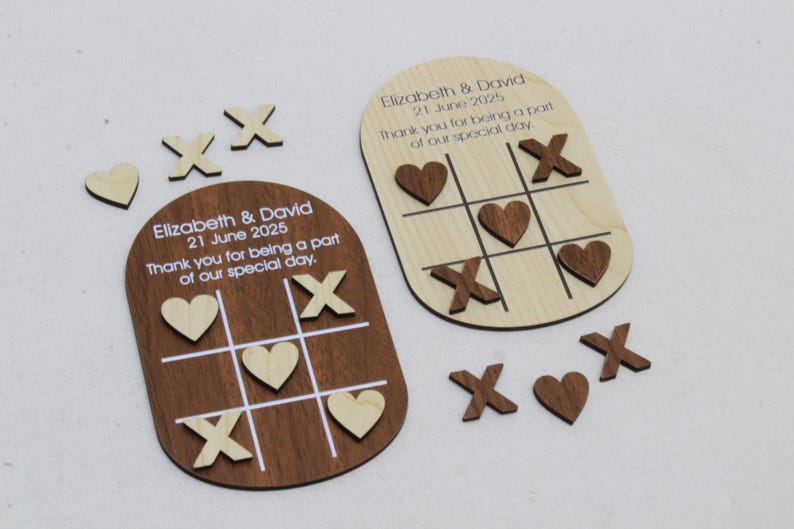 K&ouml;nnte beinhalten: Holz-Tic-Tac-Toe-Spielgeschenke mit graviertem Text. Ein Spielbrett ist dunkelbraun, das andere helles Holz. Die Spielsteine umfassen Holzherzen und X's. Der Text lautet "Elizabeth & David 21. Juni 2025".
