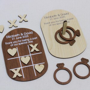 K&ouml;nnte beinhalten: Hochzeitsgeschenke aus Holz mit einem Tic-Tac-Toe-Spiel, einem Ringwurfspiel und ringf&ouml;rmigen Dekorationen. Das Tic-Tac-Toe-Spielbrett ist braun mit wei&szlig;en Linien und h&ouml;lzernen X's und Herzen. Das Ringwurfspiel befindet sich auf einer hellen Holzbasis. Der Text lautet "Elizabeth & David 21 June 2025".