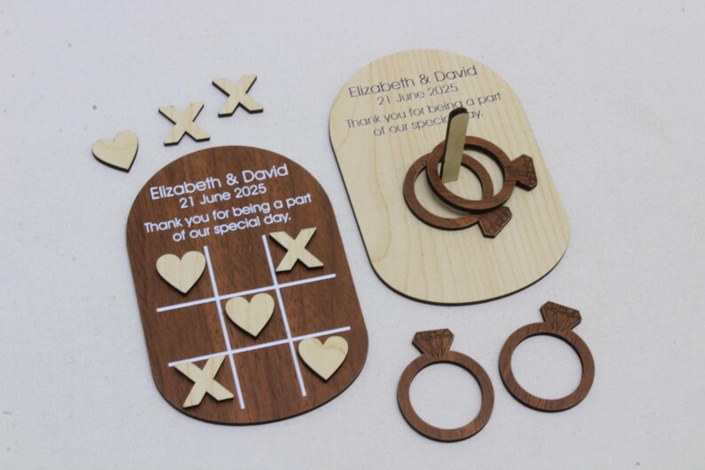 K&ouml;nnte beinhalten: Hochzeitsgeschenke aus Holz mit einem Tic-Tac-Toe-Spiel, einem Ringwurfspiel und ringf&ouml;rmigen Dekorationen. Das Tic-Tac-Toe-Spielbrett ist braun mit wei&szlig;en Linien und h&ouml;lzernen X's und Herzen. Das Ringwurfspiel befindet sich auf einer hellen Holzbasis. Der Text lautet "Elizabeth & David 21 June 2025".