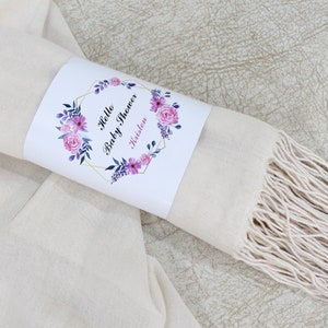 May include: A cream-colored scarf with a fringe border. The scarf is rolled up and tied with a white label that says "Hello Baby Shower Kristen" with pink and purple floral accents.