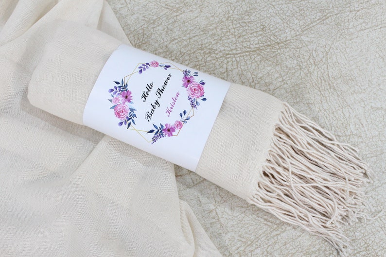 May include: A cream-colored scarf with a fringe border. The scarf is rolled up and tied with a white label that says "Hello Baby Shower Kristen" with pink and purple floral accents.
