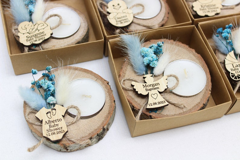 Op de afbeelding: Een set kleine houten kaarshouders met tealblauwe en witte gedroogde bloemen, een klein theelichtje en een gepersonaliseerd houten label. De labels bevatten namen en gebeurtenissen zoals "Baby Shower", "Doop" en "Baby".