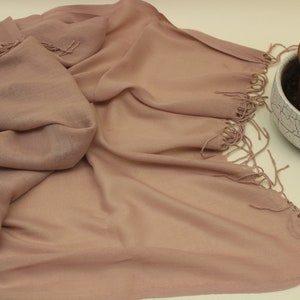May include: A soft, dusty rose colored scarf with fringed edges. The scarf is made of a lightweight fabric and is draped over a white surface.