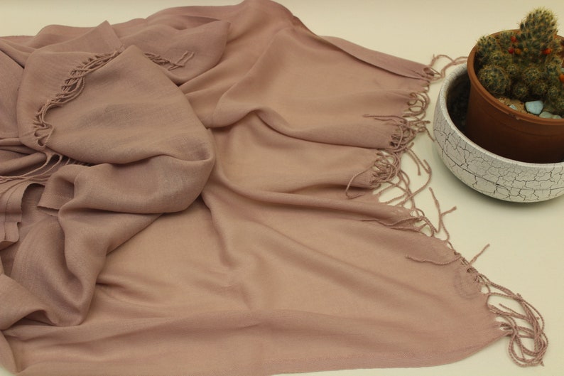 May include: A soft, dusty rose colored scarf with fringed edges. The scarf is made of a lightweight fabric and is draped over a white surface.