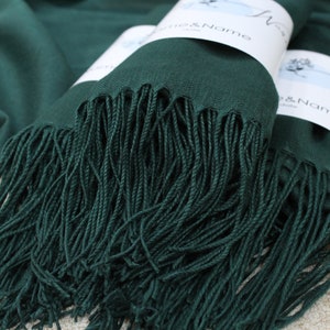May include: Two emerald green pashmina scarves with fringe, folded and stacked on a light brown surface. The scarves have white labels with the text "Woven & Name" on them.