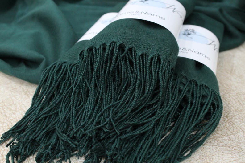 May include: Two emerald green pashmina scarves with fringe, folded and stacked on a light brown surface. The scarves have white labels with the text "Woven & Name" on them.