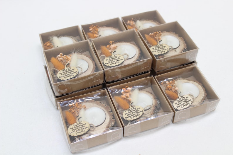 K&ouml;nnte beinhalten: Neun kleine Geschenkboxen mit transparenten Deckeln. Jede Box enth&auml;lt eine Holzscheibe mit einer wei&szlig;en Kerze, Trockenblumen und einem Etikett mit der Aufschrift "Thank You For Showering our Little Pumpkin..." f&uuml;r eine Babyparty.