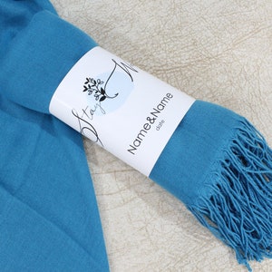 May include: A teal blue pashmina scarf with fringe, rolled up and tied with a white label that says "Stay" and "Name & Name" with the word "date" below.