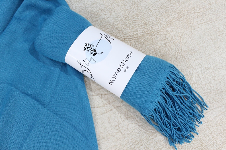 May include: A teal blue pashmina scarf with fringe, rolled up and tied with a white label that says "Stay" and "Name & Name" with the word "date" below.