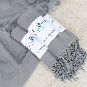 May include: Two gray scarves with fringed edges are folded and tied together with white labels that say "Stay" and "Name & Name" with a floral design.