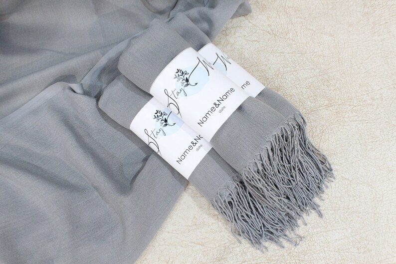 May include: Two gray scarves with fringed edges are folded and tied together with white labels that say "Stay" and "Name & Name" with a floral design.