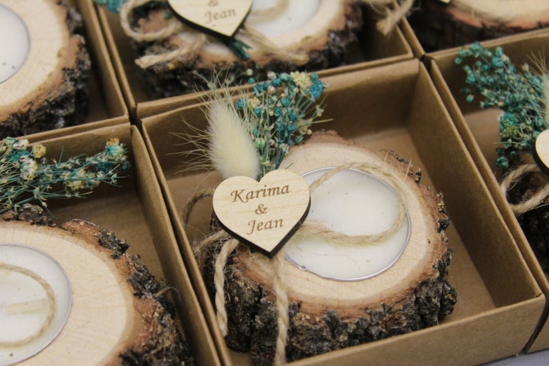 May include: A collection of rustic wooden candle holders, each crafted from a cross-section of a tree trunk. Each holder contains a white candle and is decorated with dried flowers, a fluffy accent, and a wooden heart with names engraved on it. The holders are presented in individual cardboard boxes.