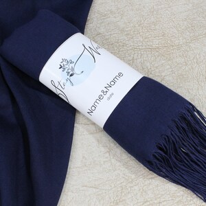 May include: A navy blue pashmina scarf with fringe, rolled up and packaged with a white label that says "Stay Wild" and "Name & Name" with the word "date" underneath.