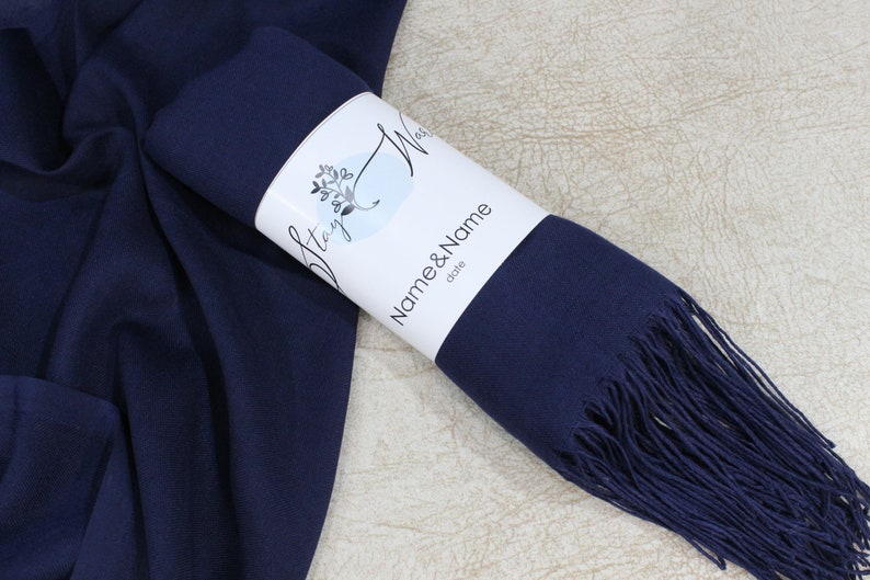 May include: A navy blue pashmina scarf with fringe, rolled up and packaged with a white label that says "Stay Wild" and "Name & Name" with the word "date" underneath.